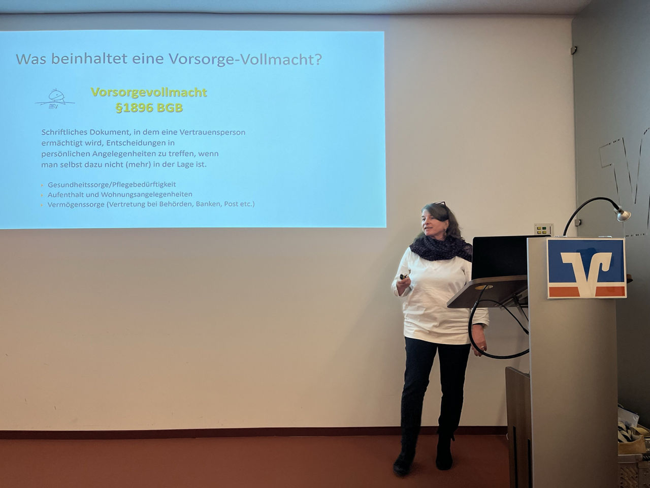 Beate Widmann - Referentin der Hospizgruppe Freising steht vor an die Wand projezierter Präsentation und spricht zum Publikum