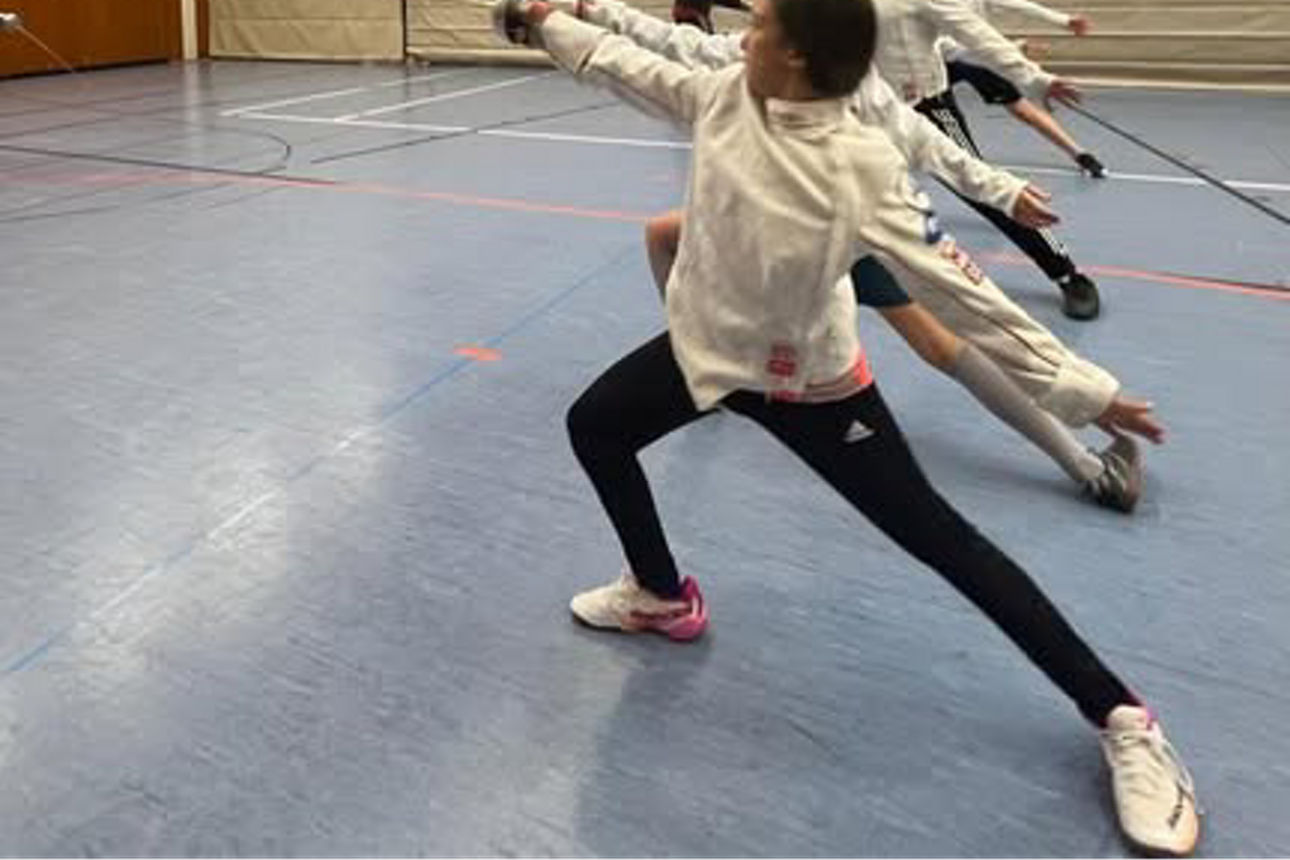 Mehrere Kinder üben gleichzeitig den Ausfall mit Degen in einer Sporthalle, angeleitet von einer Trainerin.