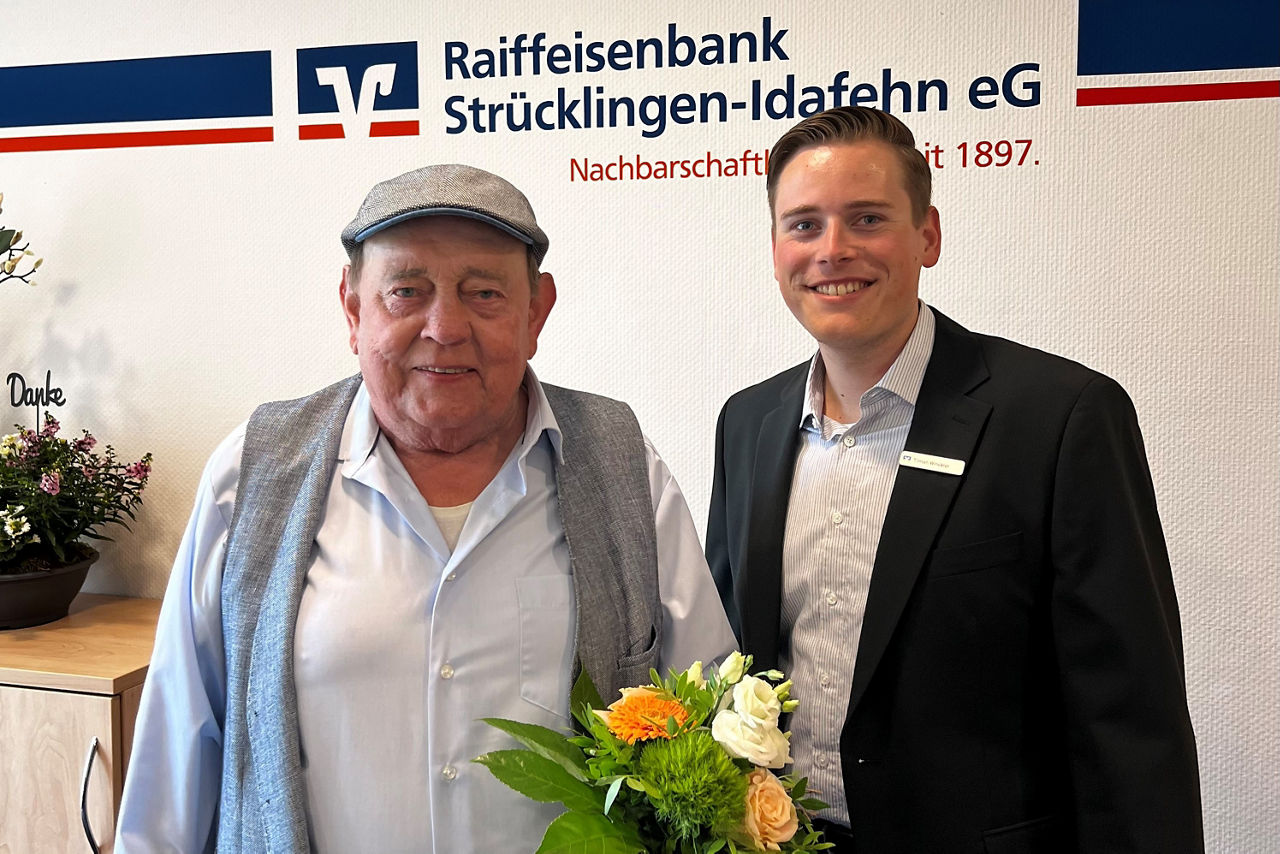 Herr Harms, Gewinner und Herr Winckler, Kundenberater stehen vor einer Wand mit dem Raiba-Logo. 