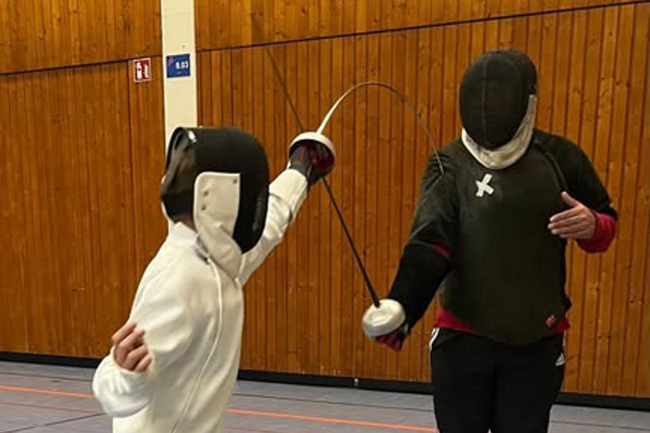 Ein Kind im Fechtanzug und mit Maske kämpft mit einem Trainer mit Maske in der Sporthalle, beide mit ausgestrecktem Degen.
