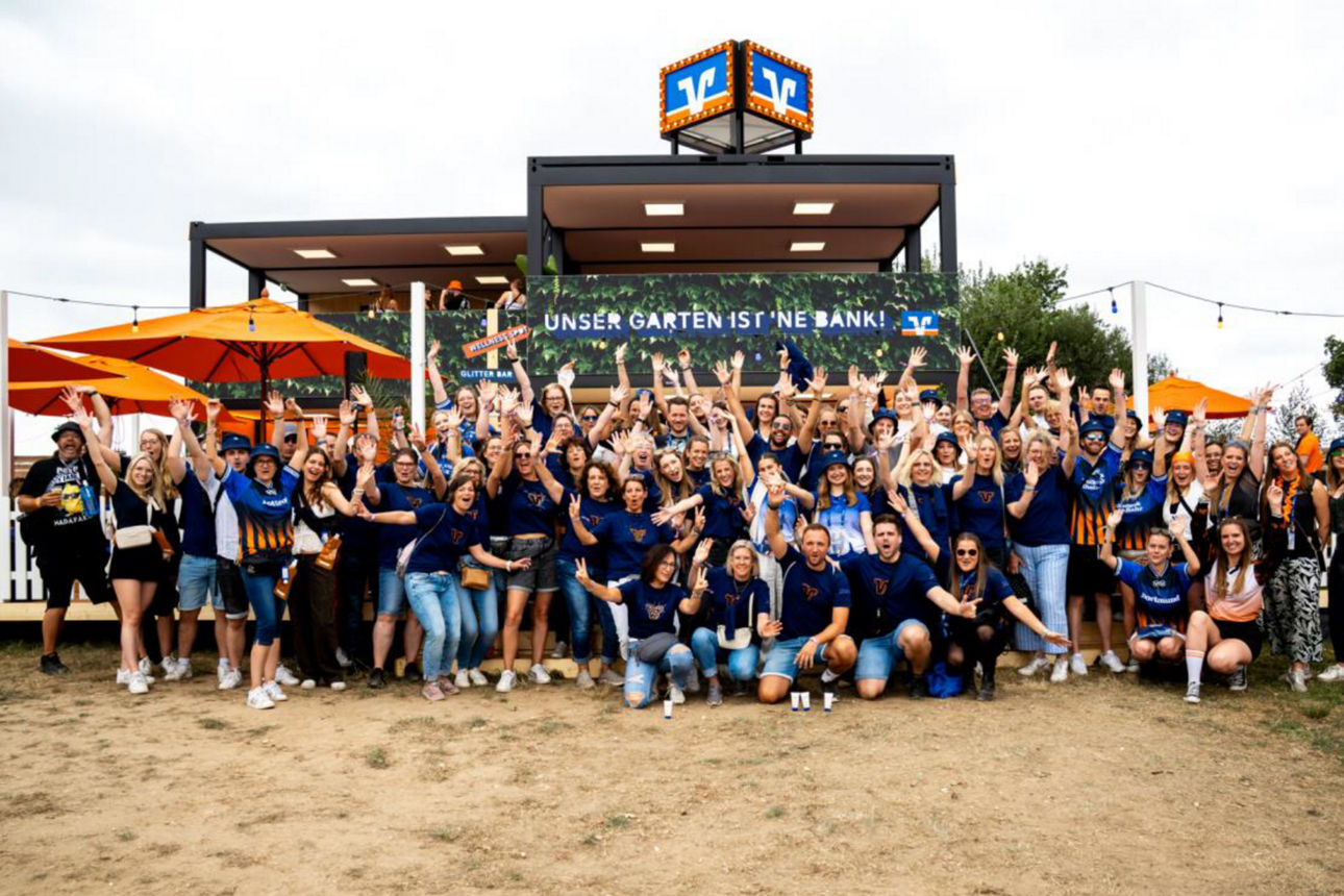 Gruppenfoto vor dem zweistöckigen Volksbank-Pavillon auf einem Festivalgelände. Rund 80 Personen stehen und knien vor dem Gebäude, viele mit erhobenen Armen. Über der Terrasse des Pavillons ist ein großes Banner mit dem Schriftzug „UNSER GARTEN IST ’NE BANK!“ angebracht, auf dem Dach befindet sich ein beleuchtetes Volksbank-Logo. Links und rechts des Pavillons stehen mehrere orangefarbene Sonnenschirme, Lichterketten und weiße Zaunelemente begrenzen die Fläche. Das Foto zeigt die gemeinschaftliche Atmosphäre einer Veranstaltung im Freien auf einem weitläufigen Gelände. Unten mittig auf dem Bild ist ein schwarzes Rechteck mit weißem Schriftzug "Teilnahmebedingungen San Hejmo".