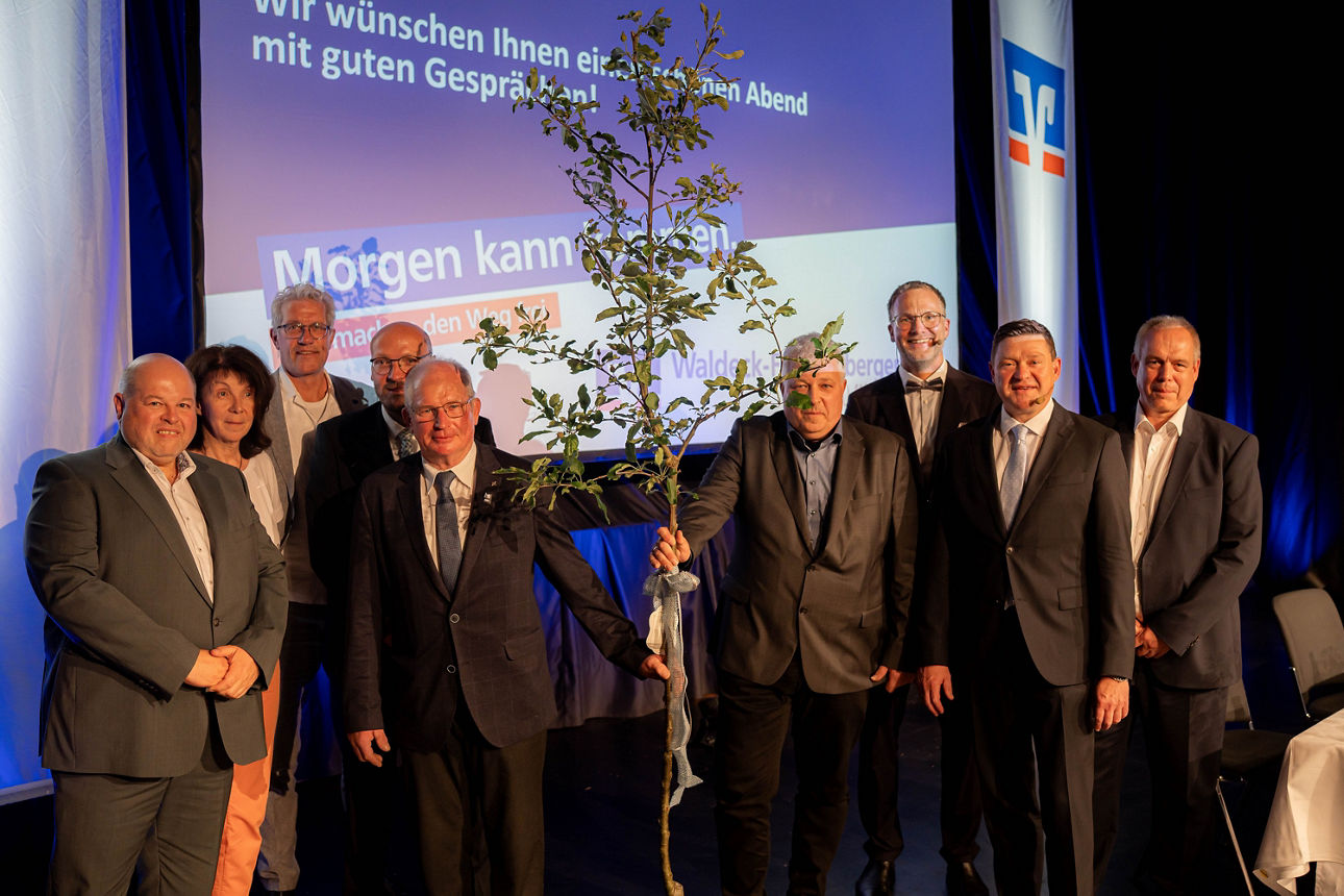Gruppe von Personen bei Veranstaltung mit symbolischer Baumpflanzung, gemeinsames Foto auf Bühne vor Präsentationswand.