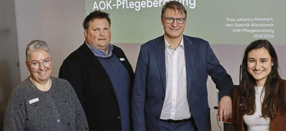 Zwei Frauen und zwei Männer (mittig) stehen nebeneinander und lächeln in die Kamera. Im Hintergrund ein Ausschnitt  einer Präsenttion an der Wand mit dem Text: AOK-Pflegeberatung