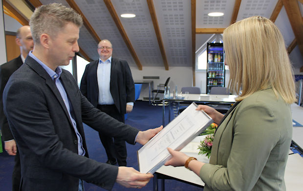 Ein Mann in einem dunkelblauen Anzug und einem hellblauen Hemd überreicht einer Frau in einem hellgrünen Blazer eine gerahmte