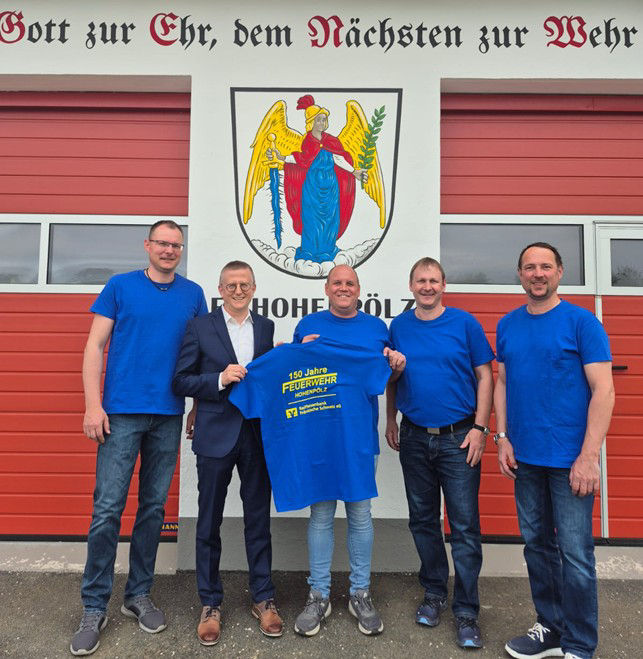 Gruppe vor Feuerwehrhaus präsentiert T-Shirt bei gemeinsamer Veranstaltung.