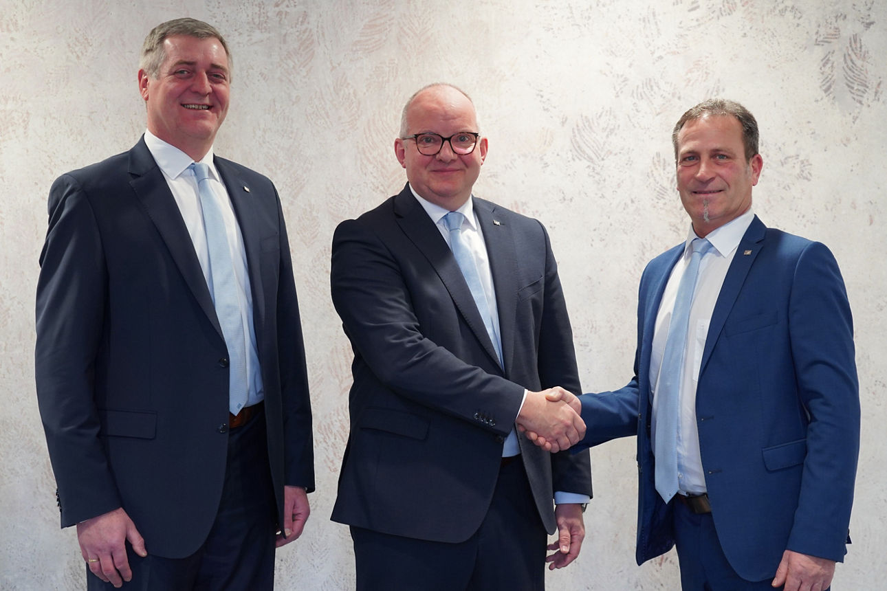 Drei Männern in Anzügen lächeln in die Kamera. Zwei Männer schütteln sich dabei die Hände