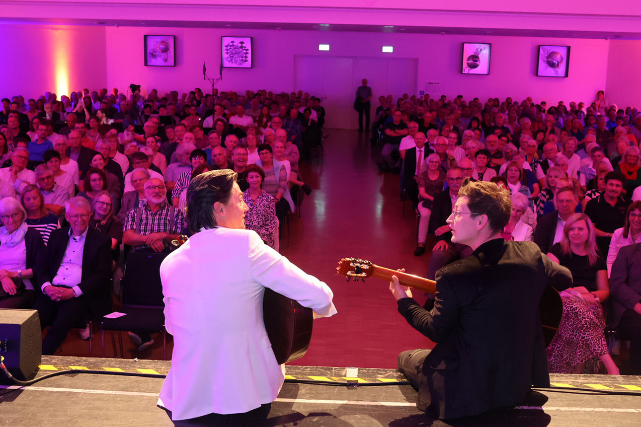Zwei Männer sitzen auf einer Bühne und spielen Gitarre vor vielen Menschen