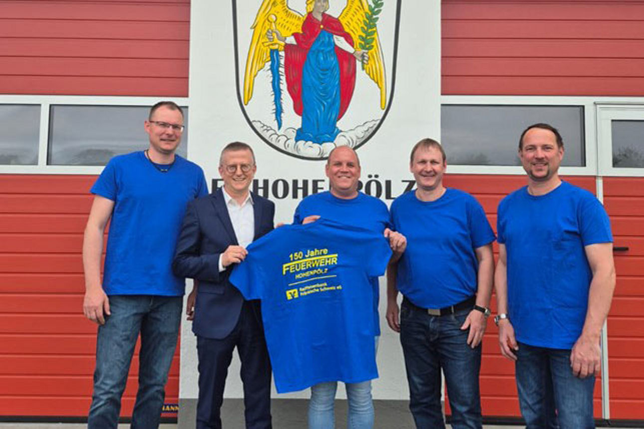 Gruppe vor Feuerwehrhaus präsentiert T-Shirt bei gemeinsamer Veranstaltung.