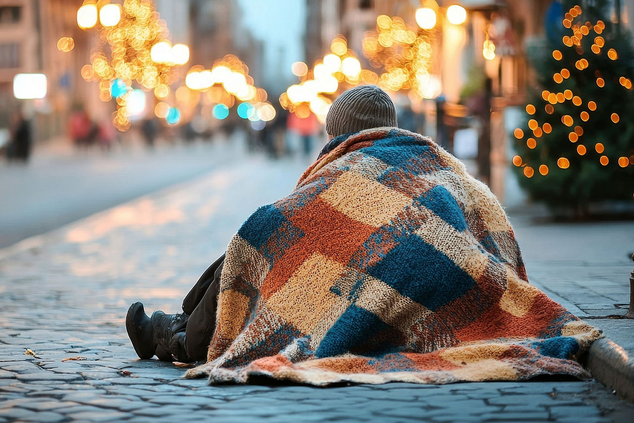Ein Wohnunsloser sitz auf Kopfsteinpflaster zur Weihnachtszeit in der Stadt mit Mütze und Decke