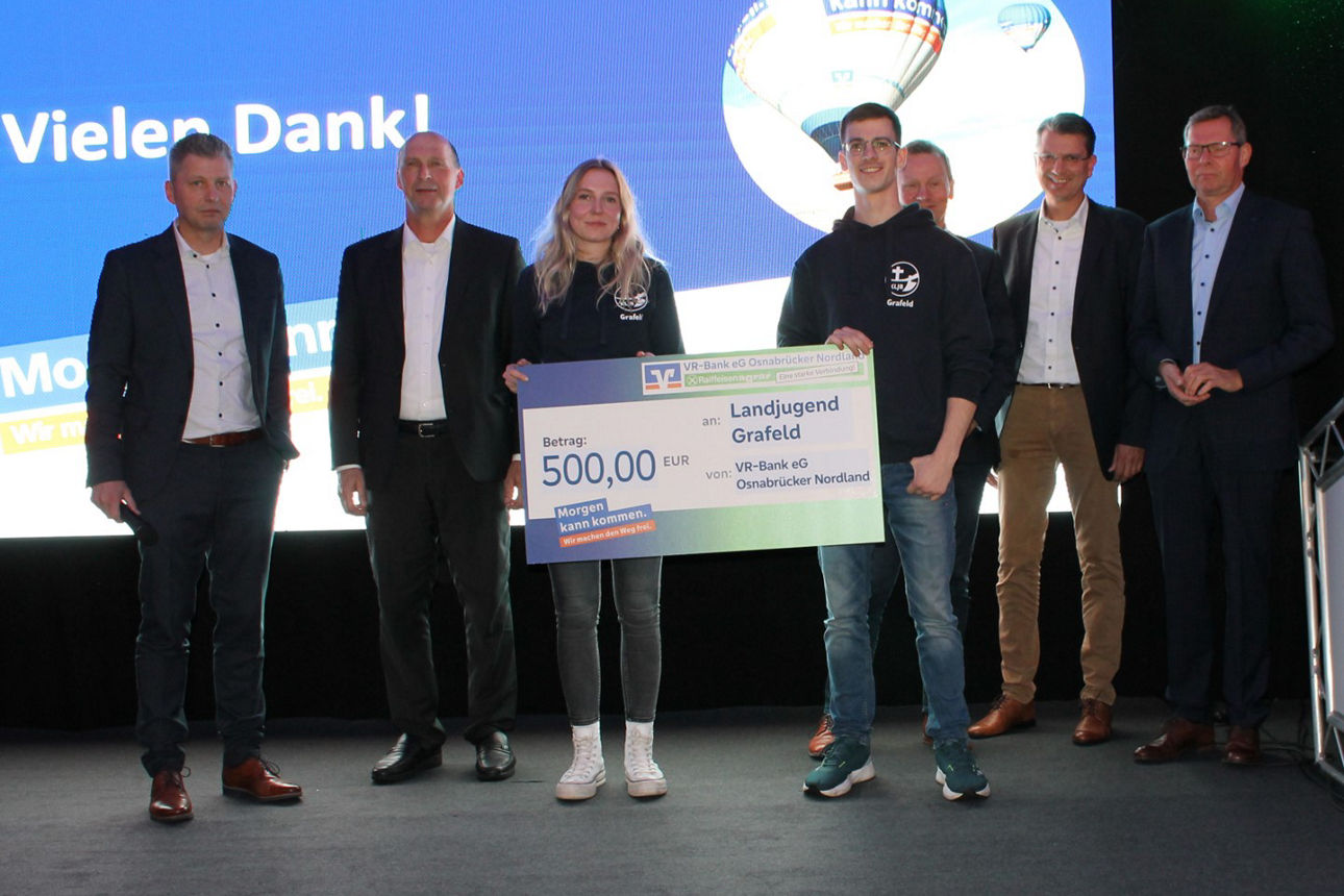 Eine Gruppe von Menschen steht vor einem großen Bildschirm, auf dem ein Scheck angezeigt wird, und diskutiert oder jubelt.