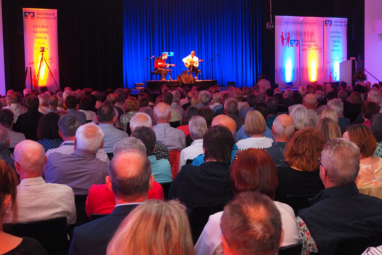 Zwei Männer sitzen in einem vollbesetzten Saal auf der Bühne und spielen Gitarre