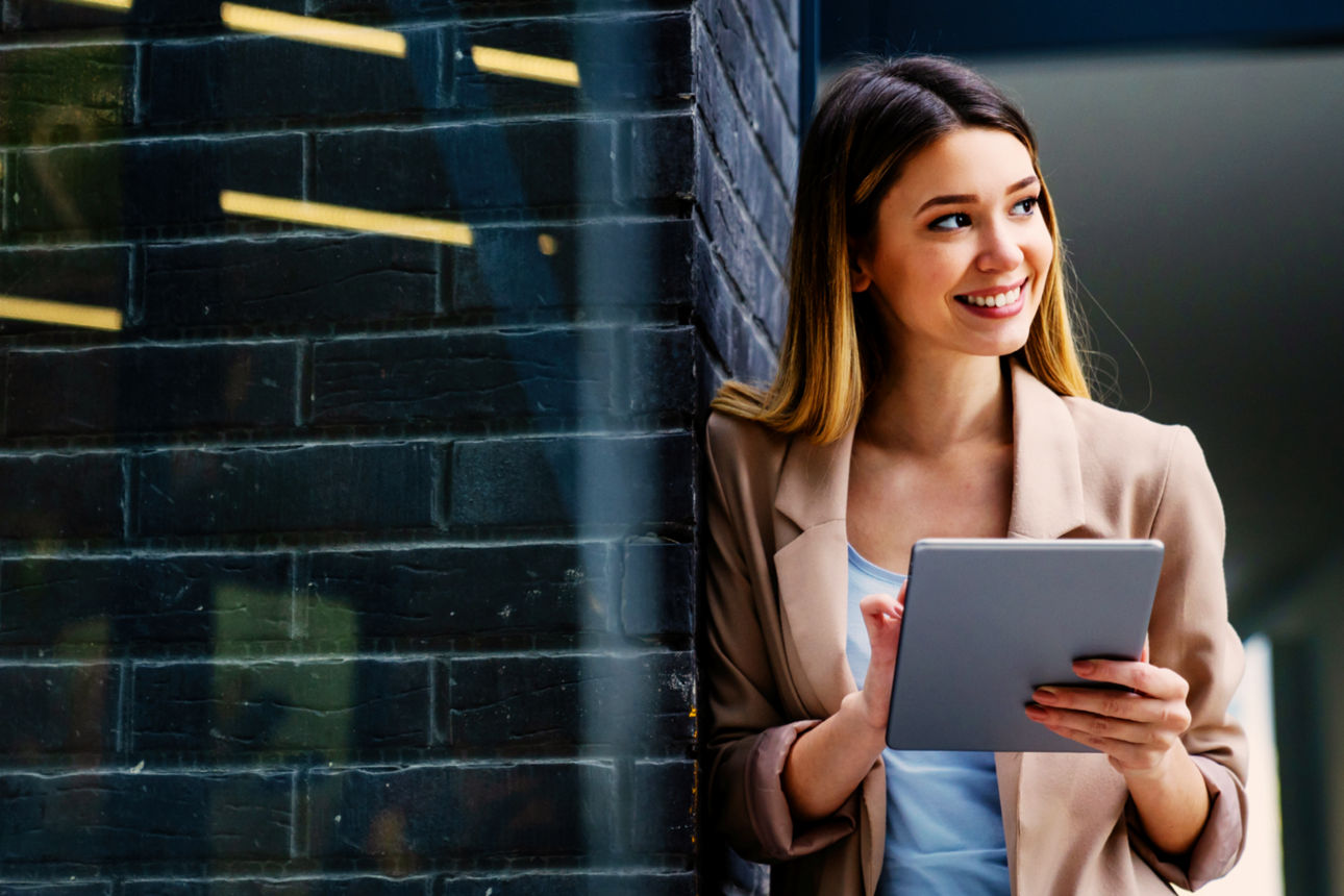 Frau stehtl lächelnd an einer Wand und hat ein Tablet in der Hand
