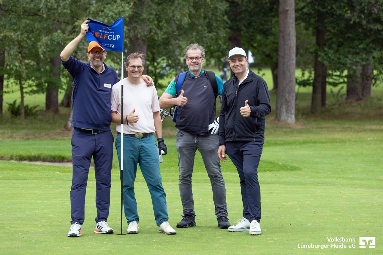 Ein Flight mit vier gut gelaunten Personen steht an einem Loch auf dem Golfplatz, einer hält die Volksbankfahne nach oben.