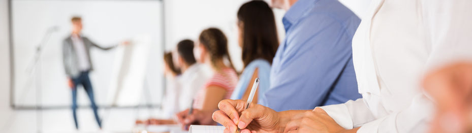 Teilnehmende einer Schulung sitzen in Reihe und machen Notizen, während eine Person vorne präsentiert