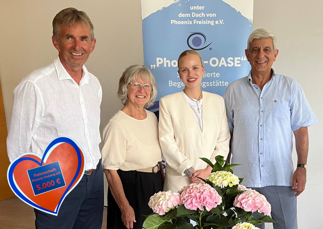 Vier Menschen stehen vor einer Wand mit dem Vereinslogo von Phoenix Freising e.V. und schauen in die Kamera; Im Vordergrund eine rosa Hortenisie und links ein Spendenherz mit dem Betrag  Euro