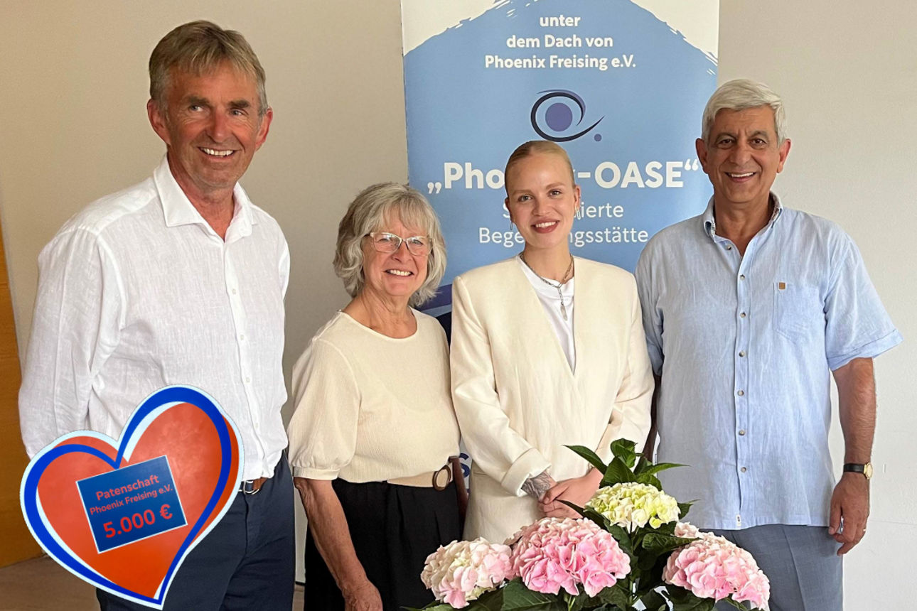Vier Menschen - rechts und links je ein Mann, in der Mitte zwei Frauen - stehen vor einer Wand mit dem Vereinslogo von Phoenix Freising e.V. und schauen in die Kamera; Im Vordergrund eine rosa Hortenisie und links ein Spendenherz mit dem Betrag  Euro