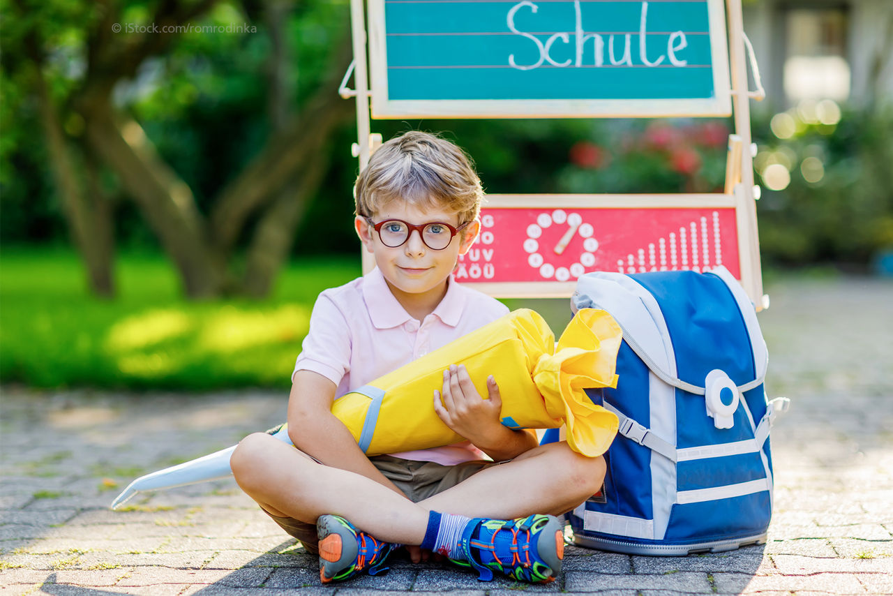 Kind mit Schultüte und Ranzen sitzt auf dem Boden draußen vor einer kleinen Tafel