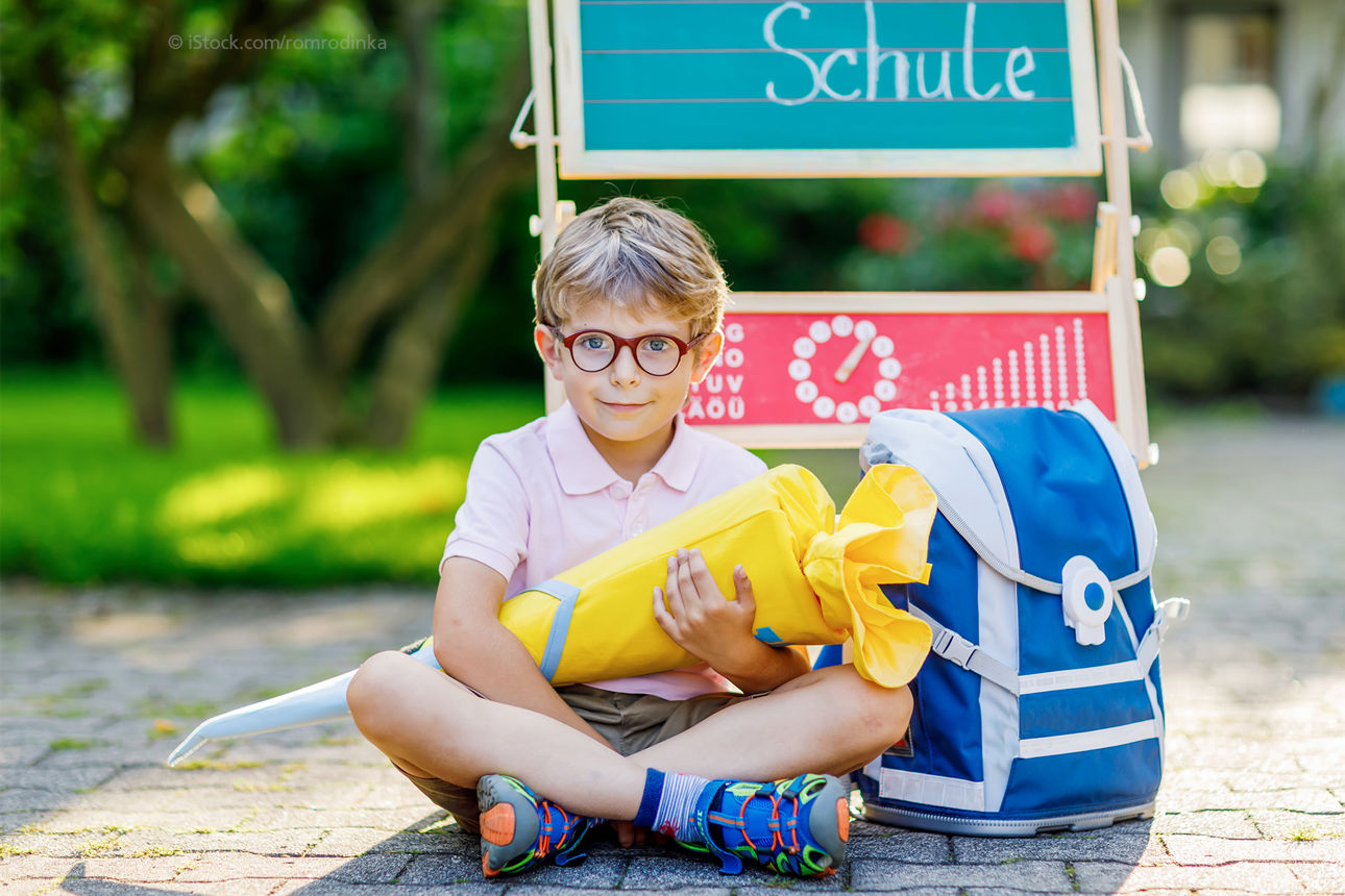 Kind mit Schultüte und Ranzen sitzt auf dem Boden draußen vor einer kleinen Tafel