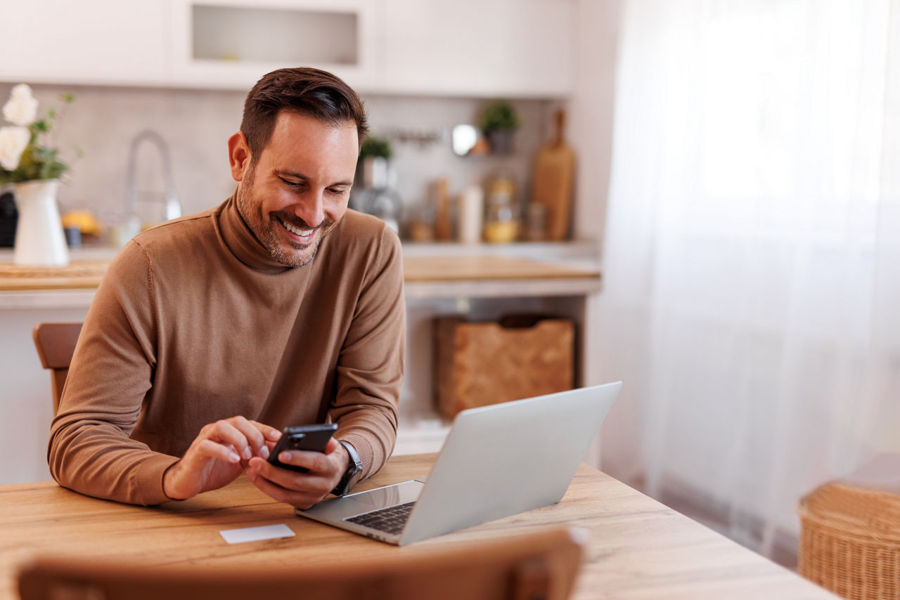 Porträt eines lächelnden Mannes mittleren Alters mit Kreditkarte, der zu Hause auf dem Esstisch Mobiltelefon und Laptop benutzt