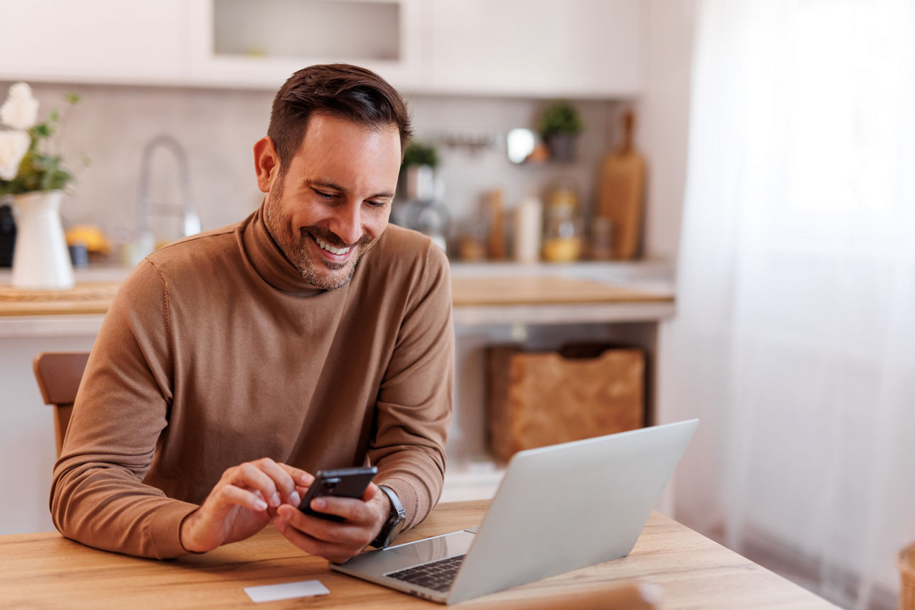 Porträt eines lächelnden Mannes mittleren Alters mit Kreditkarte, der zu Hause auf dem Esstisch Mobiltelefon und Laptop benutzt