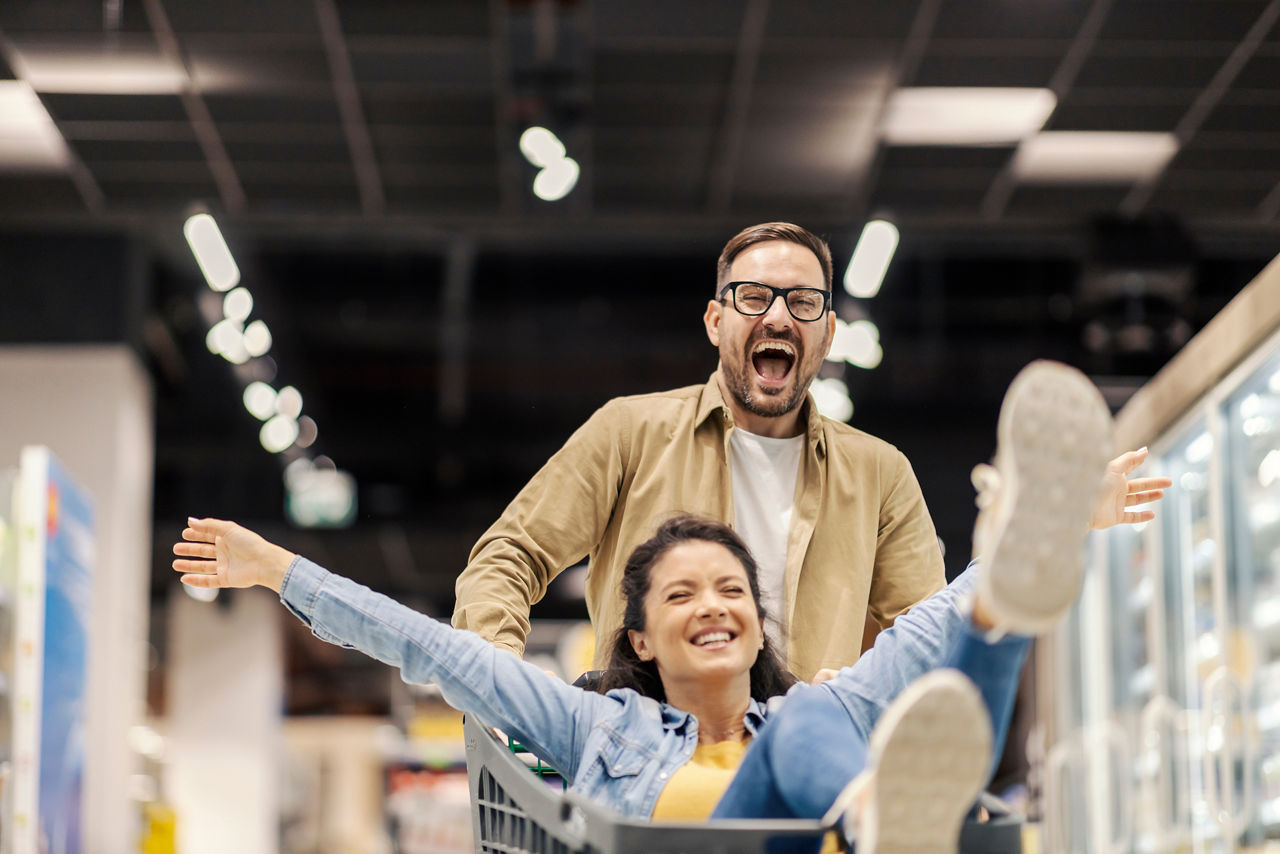 Ein fröhlicher Mann schiebt eine Frau im Einkaufswagen im Supermarkt.