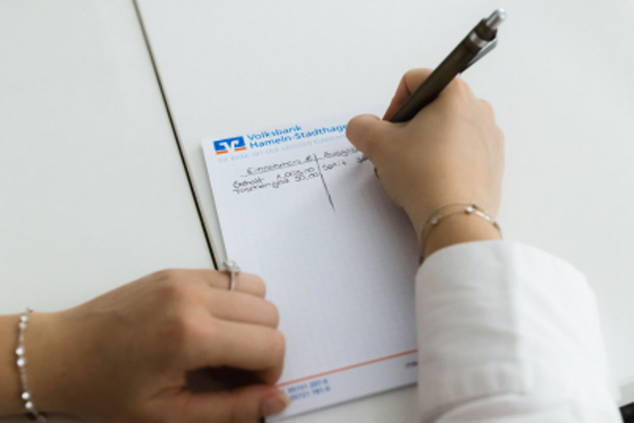 Eine Person schreibt auf einem Notizblock der Volksbank Hameln-Stadthagen handschriftliche Notizen an einem Schreibtisch.
