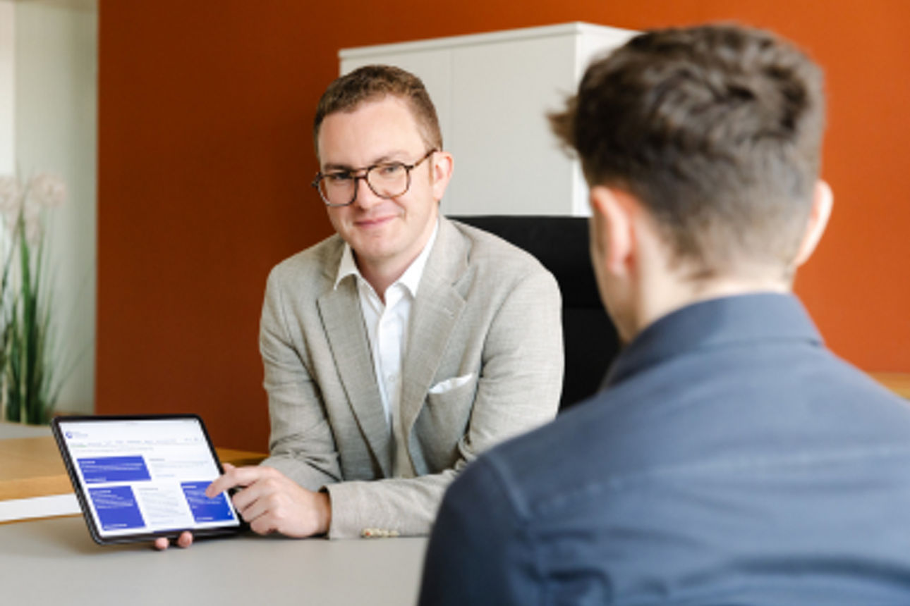 Zwei Personen in einem Büro, wobei eine Person auf einem Tablet Informationen präsentiert und diese der anderen erklärt.