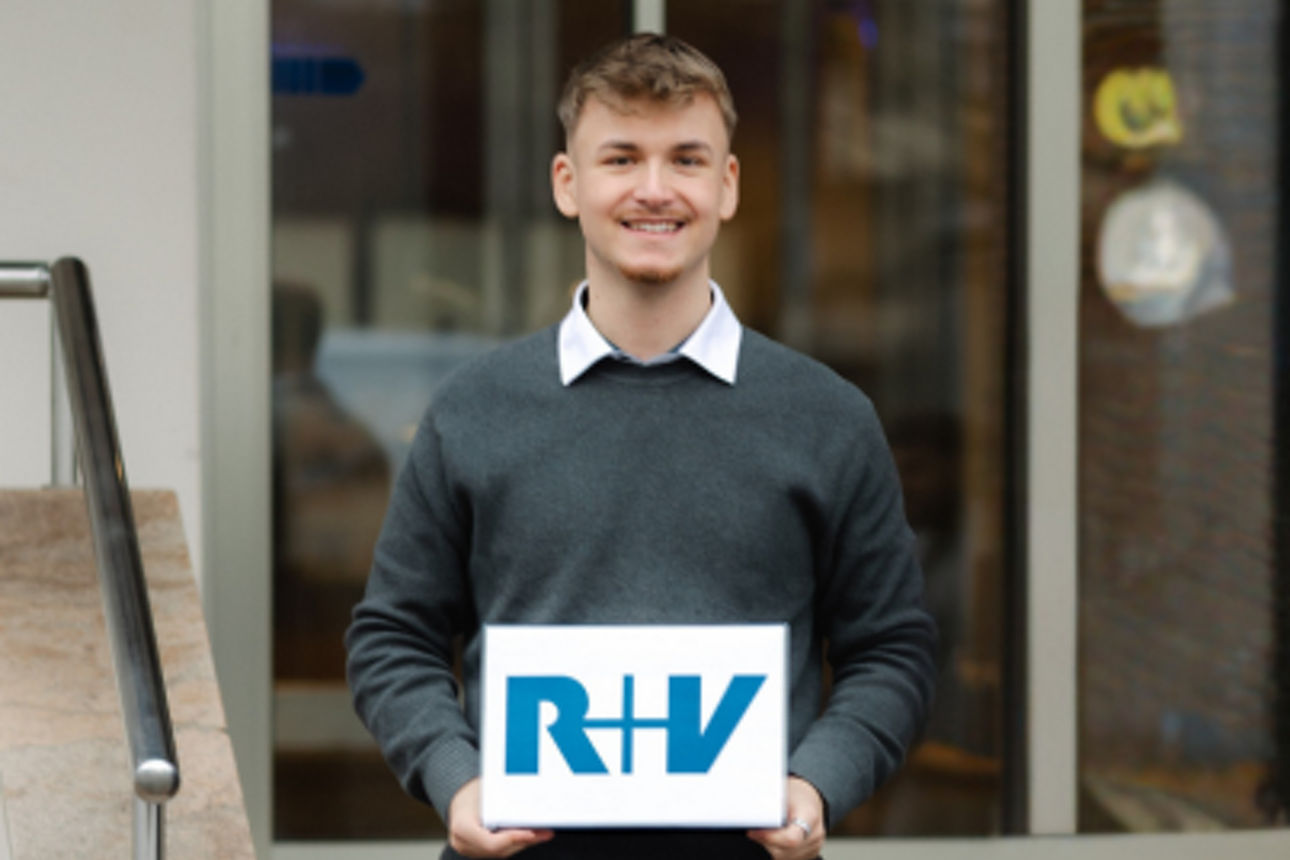 Eine Person steht vor einem Gebäude und hält ein Schild mit dem Logo der R+V Versicherung in den Händen.