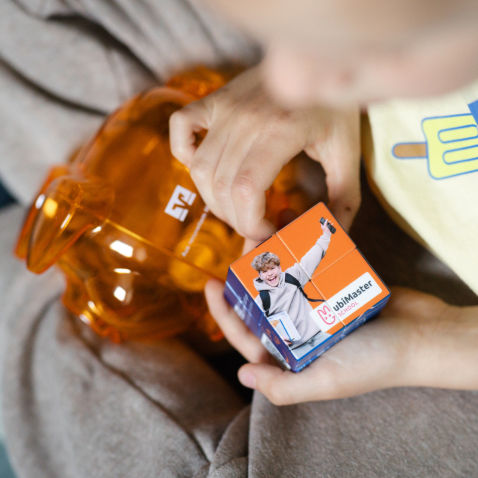 Ein Kind hat ein orangefarbenes Sparschwein im Schoß und hält einen Zauberwürfel in der Hand.
