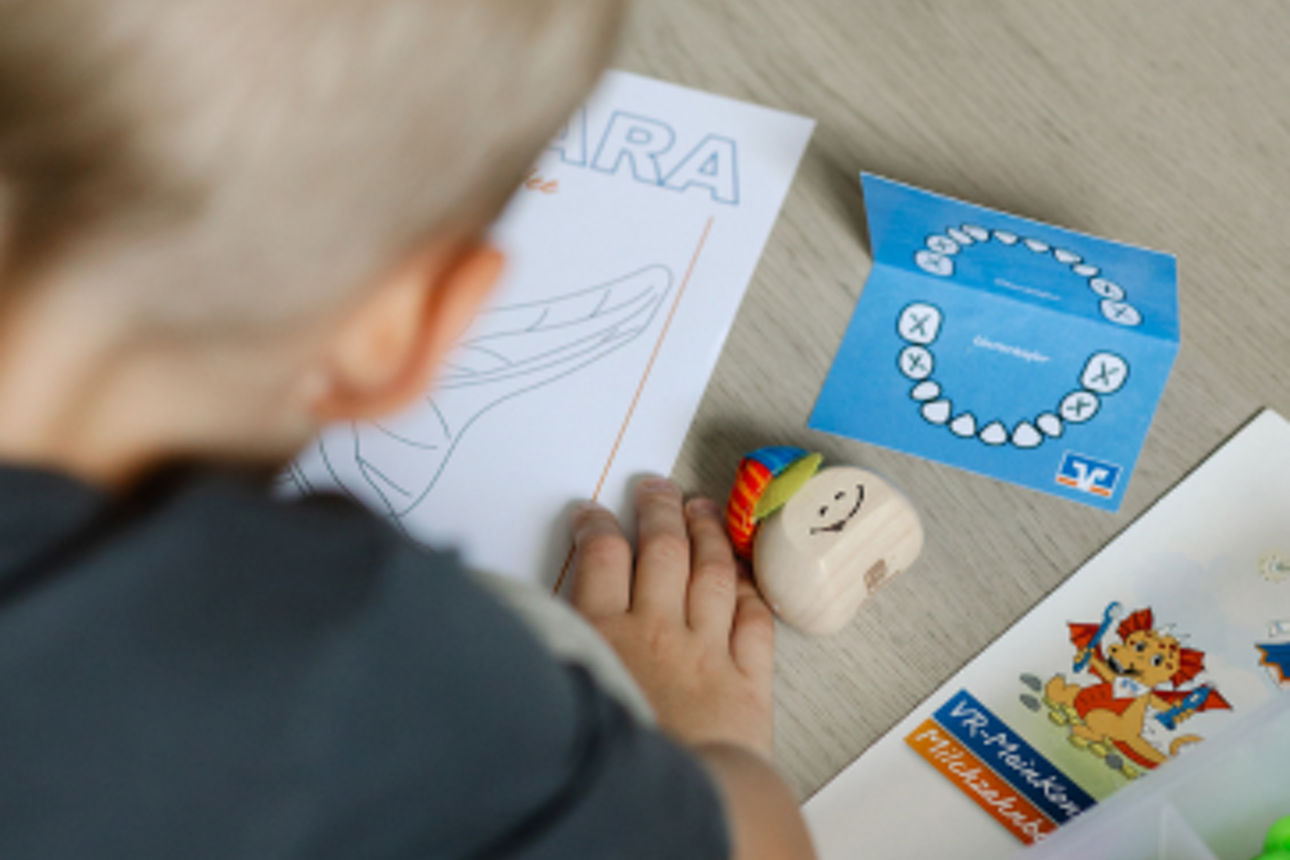 Ein Kind beschäftigt sich mit Unterlagen und Spielmaterial auf dem Tisch, daneben liegt eine Milchzahnkarte und -dose.