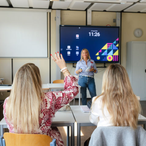 Eine Schulungs- oder Unterrichtssituation mit Teilnehmenden an Tischen und einer Präsentation auf einem großen Bildschirm.