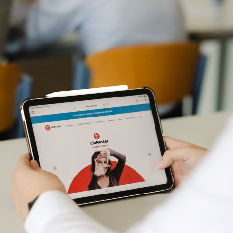 Auf einem Tablet wird die Lernplattform ubiMaster dargestellt, während das Gerät in einem Unterrichtsraum genutzt wird.