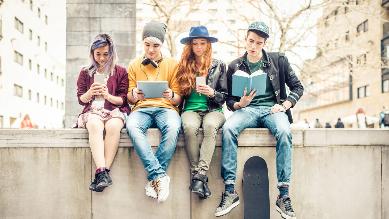 4 Jugendliche sitzen auf einer Mauer und lesen Bücher und an ihrem Smartphone