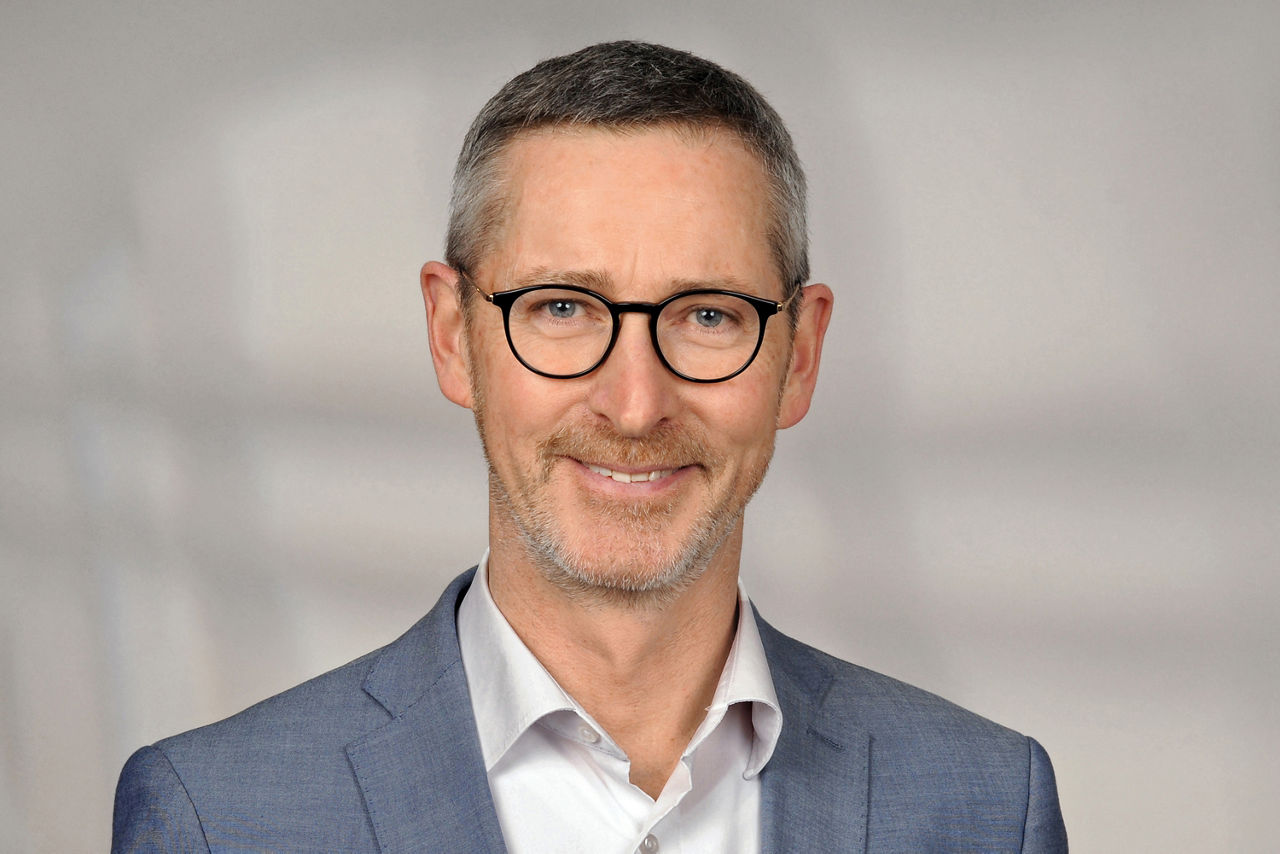 Andreas Boos hat kurze graue Haare und trägt eine schwarze halbrunde Brille und einen Anzug in blaugrau.