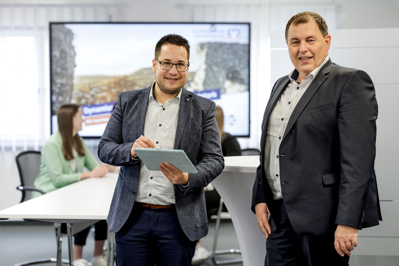 Bankberater steht vor einem großem Bildschirm und hält ein blaues Tablet in der Hand.