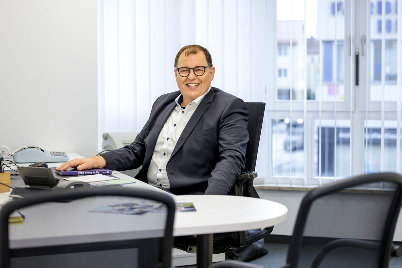 Bankberater mit Anzug sitz am Bürotisch im Büro und schaut in die Kamera.