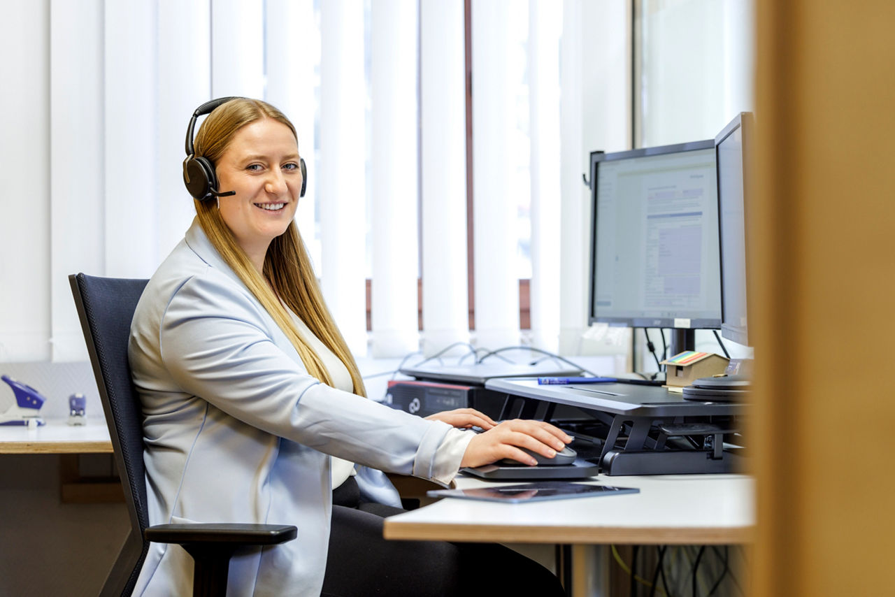Eine Frau sitz am PC mit Headset und telefoniert und blickt hierbei in die Kamera