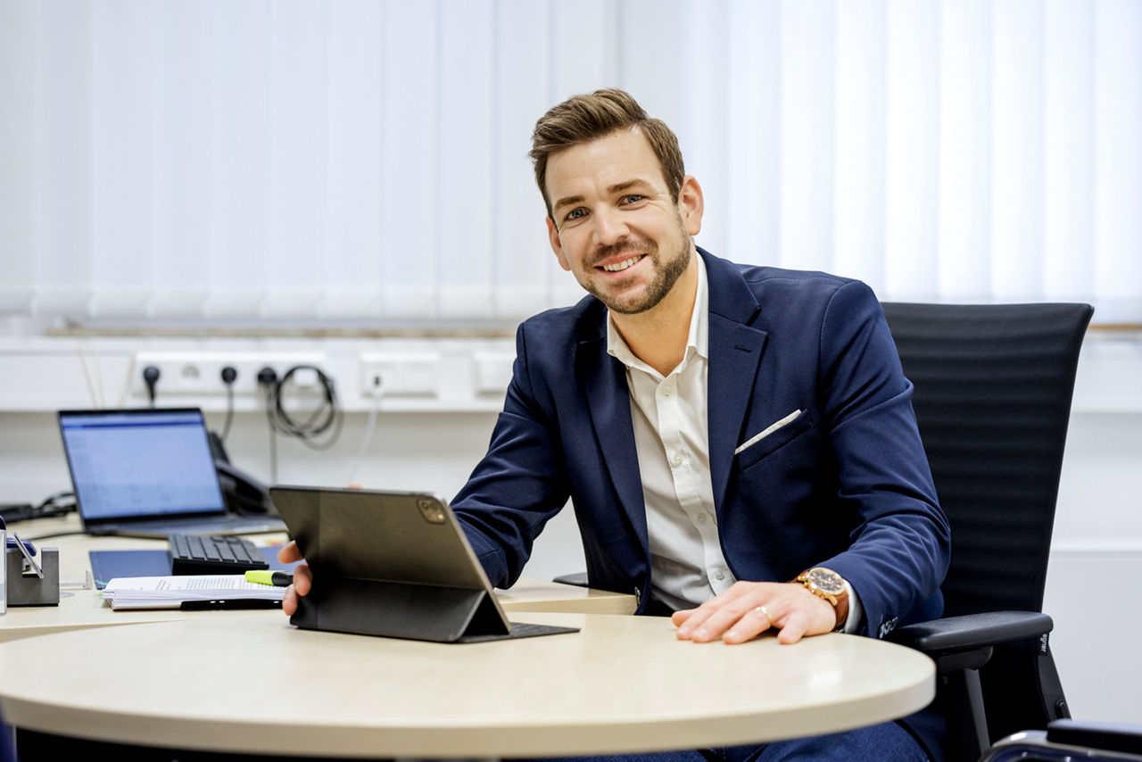 EIn Berater sitzt am Bürotisch mit einem Tablet und lächelt in die Kamera.