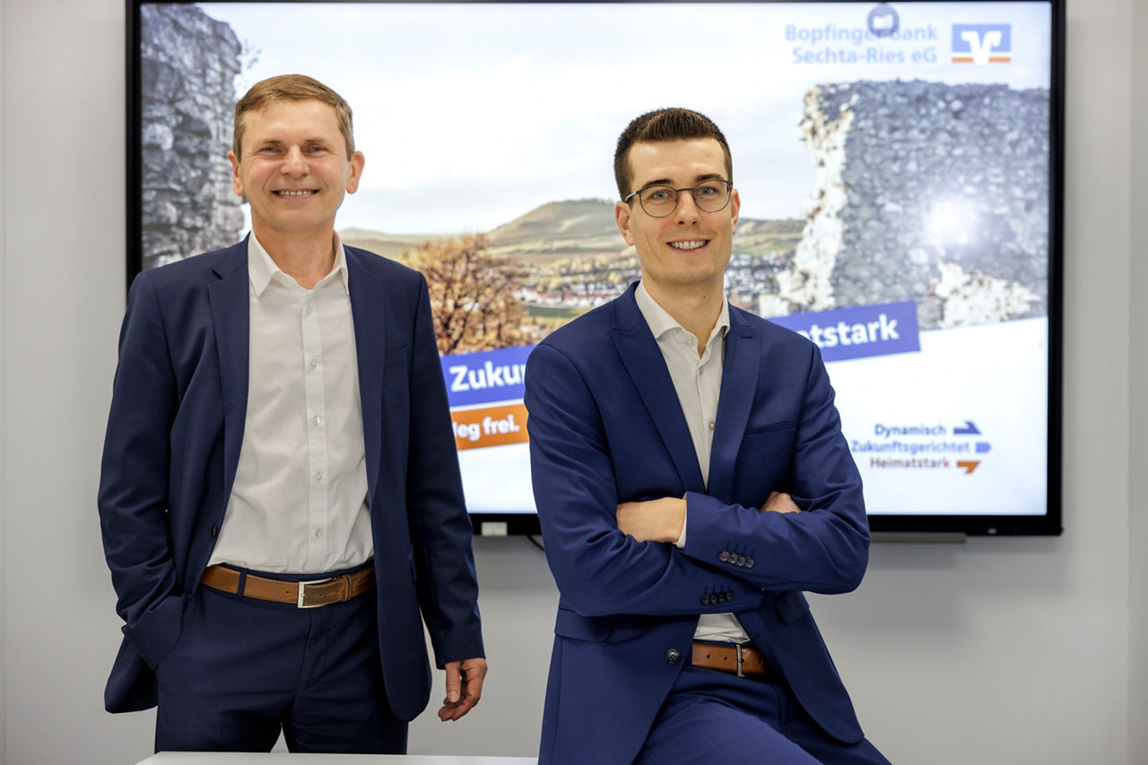 Zwei männliche Vorstände stehen vor einem Bildschirm mit einem regionalen Bild und schauen in die Kamera