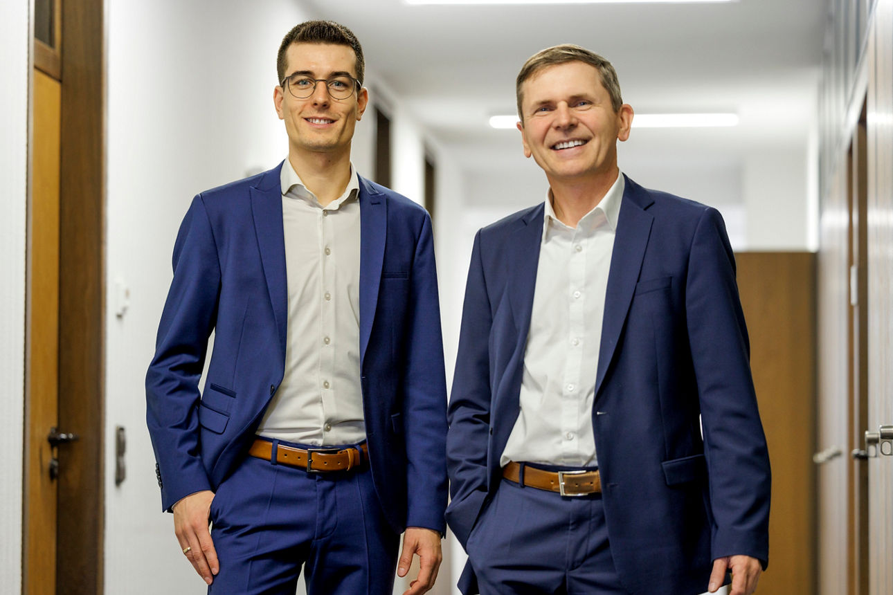 Zwei männliche Vorstände in Anzügen stehen in einem Flur, beide schauen freundlich in die Kamera.
