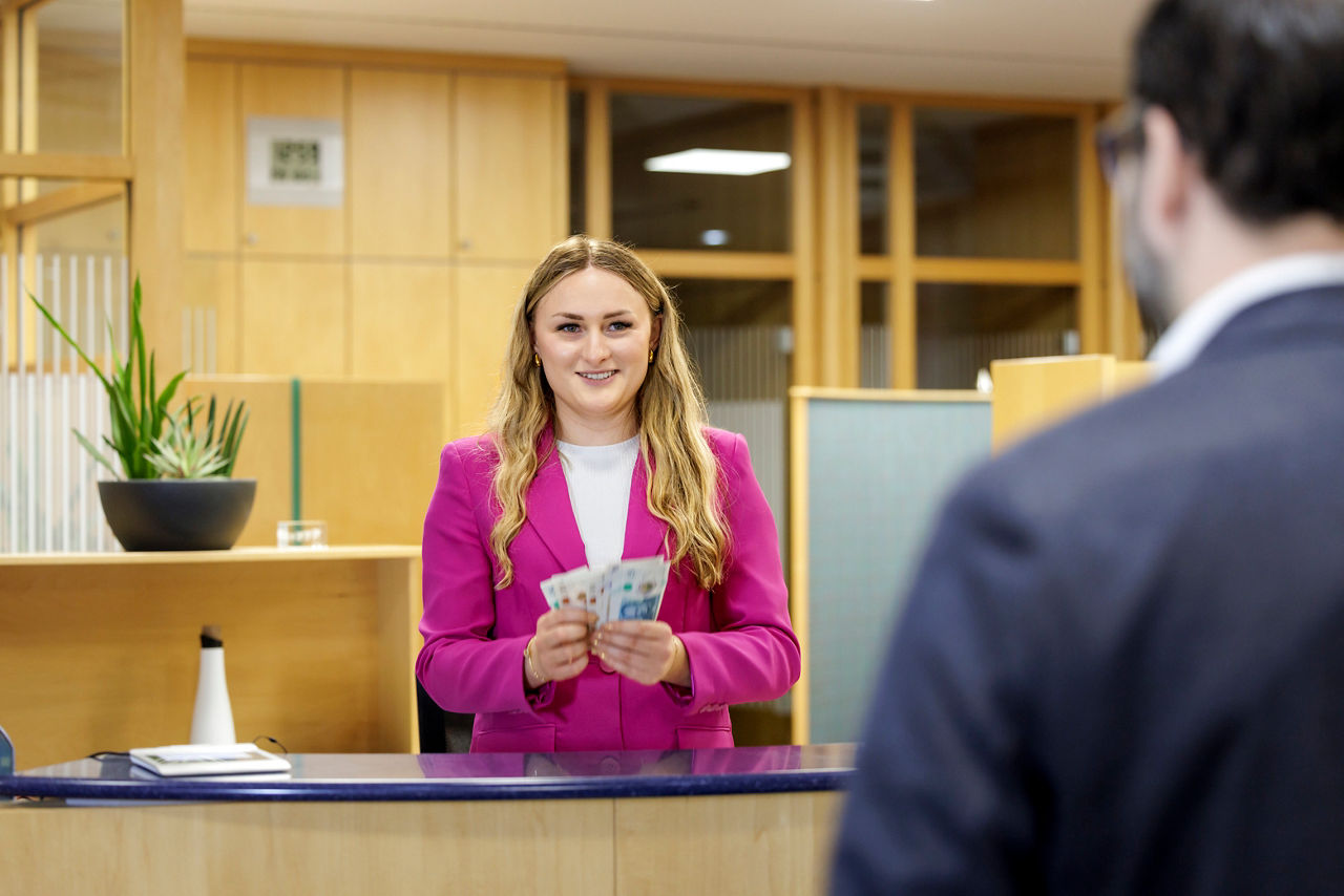 Eine Beraterin in der Bank steht am Schalter und zahlt einem Kunden Bargeld aus. 