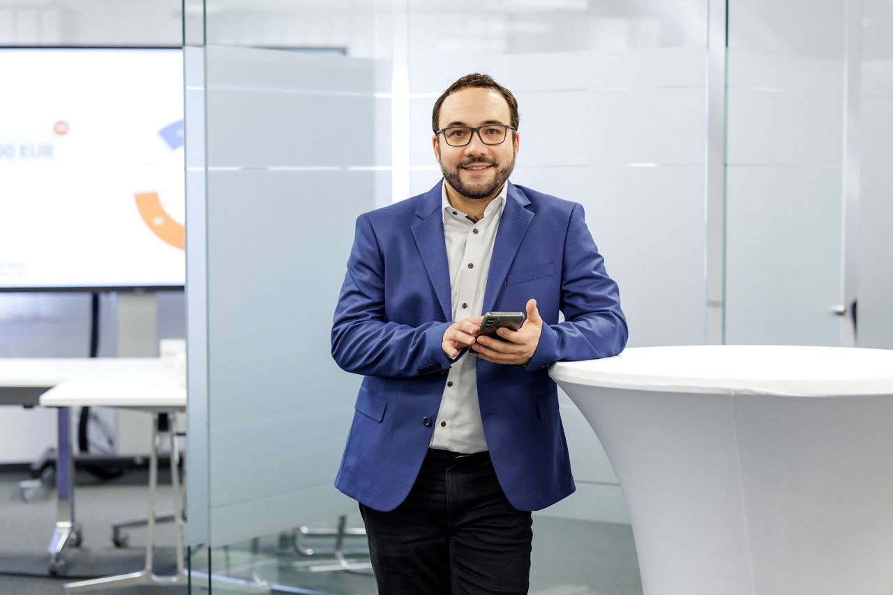 Bankberater mit Anzug steht an einem Stehtisch, hat ein Smartphone in der Hand und schaut freundlich in die Kamera.