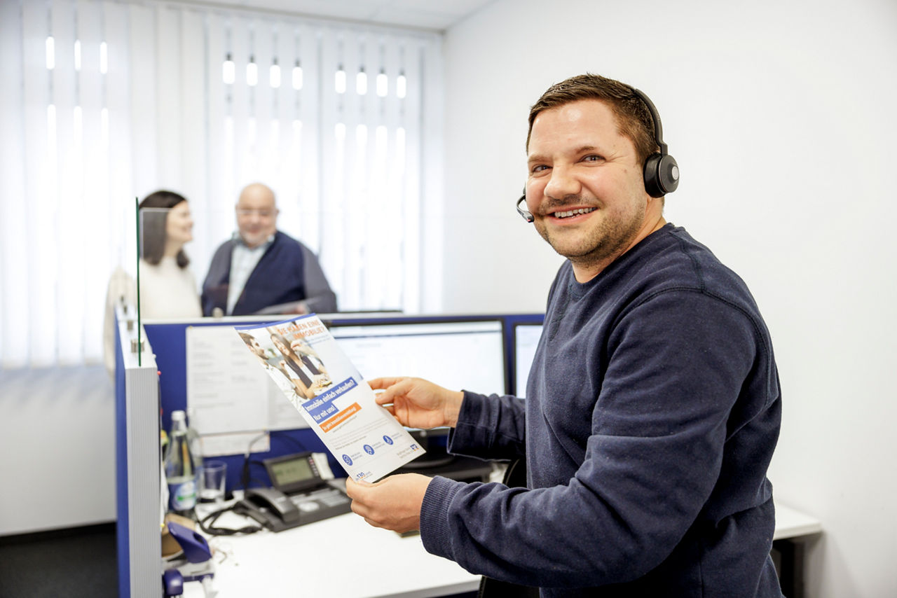 Ein Mann mit Headset und einem Flyer steht im Büro und telefoniert, im Hintergrund ist eine Frau und ein Mann zu sehen.