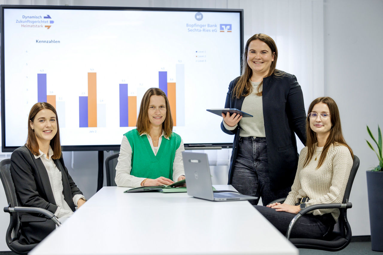 Vier Frauen sitzen am Tisch mit einem Laptop und einem Tablet und schauen freundlich in die Kamera