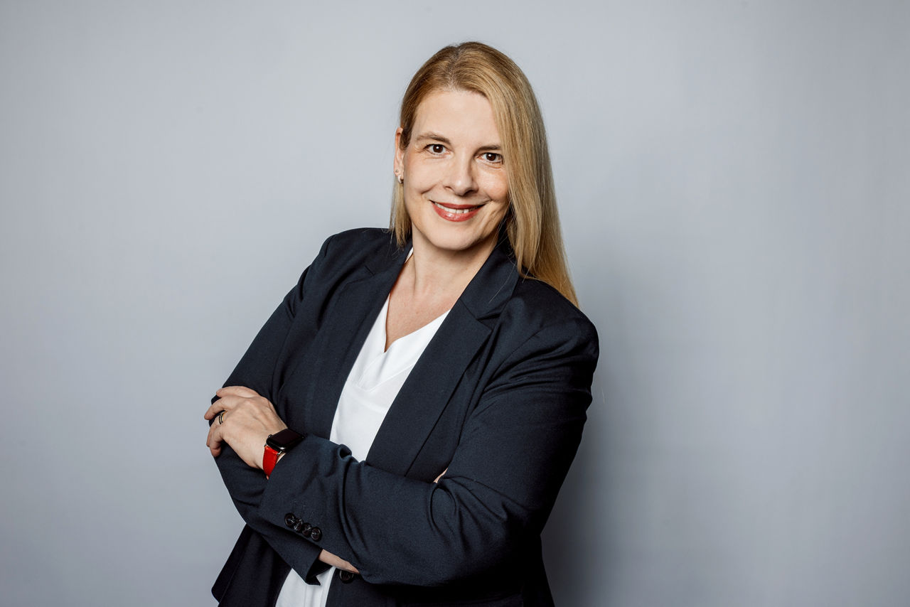 Eine Blonde Dame mit einem Blazer vor einem grauen Hintergrund.