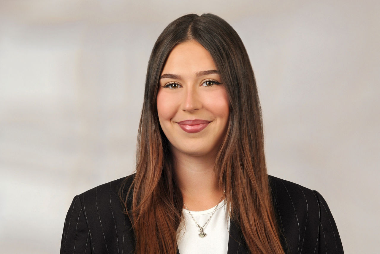 Frau mit langen braunen Haaren trägt schwarzen Nadelstreifenblazer und weiße Bluse, Halskette mit Anhänger