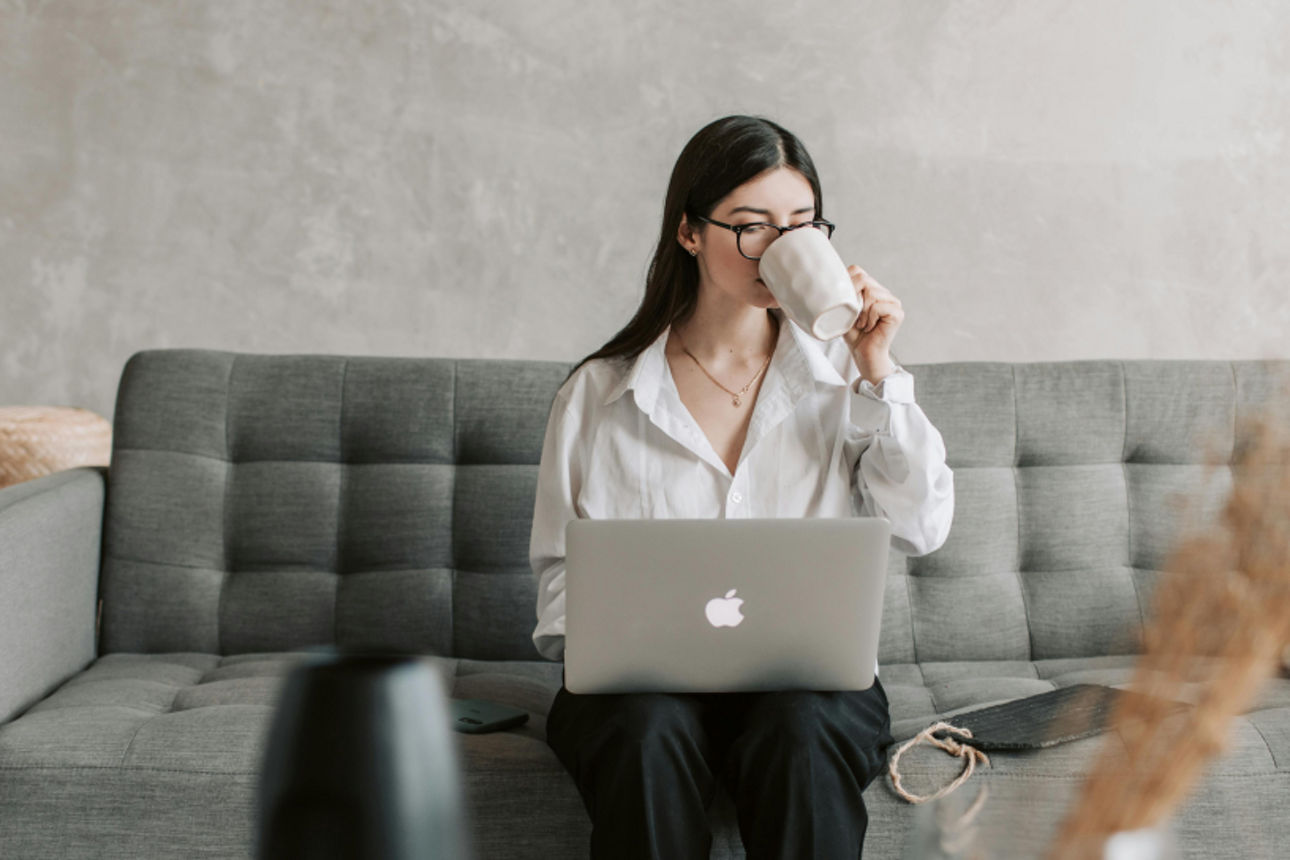 Frau sitzt auf einer Couch, trinkt einen Kaffee und hat einen Laptop auf dem Schoß