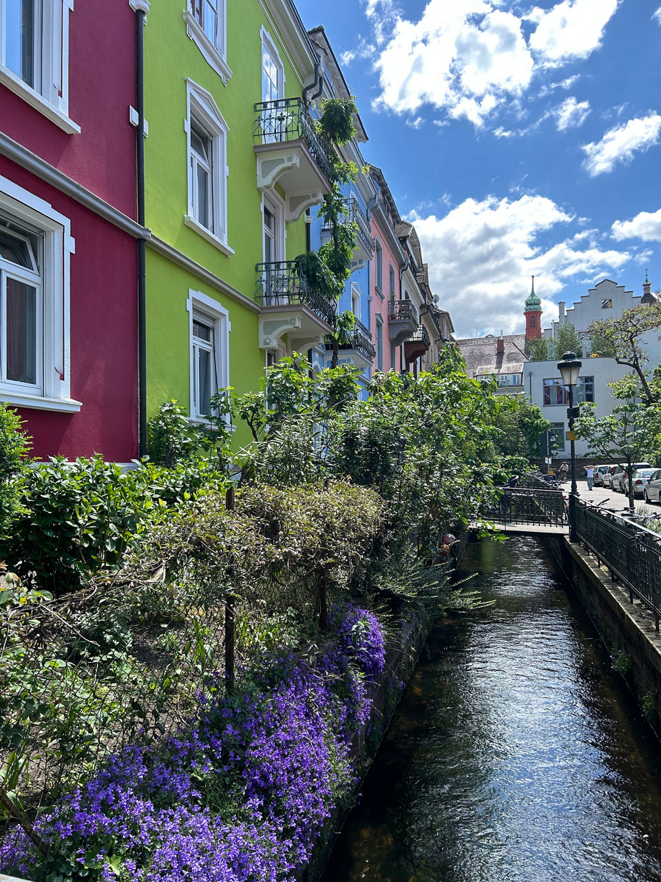 Ein schmaler Bach fließt zwischen farbenfrohen Häusern und blühenden Pflanzen unter blauem Himmel hindurch.