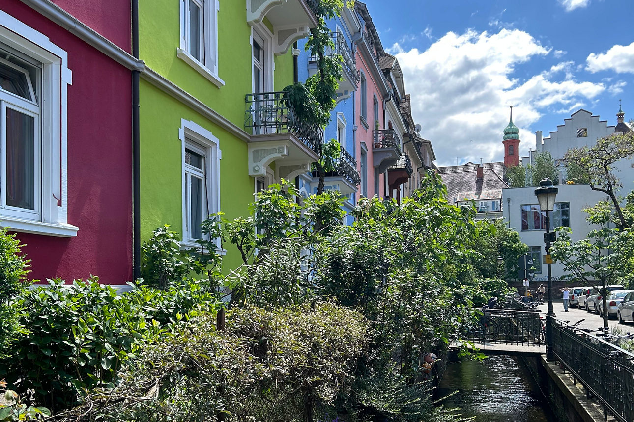 Ein schmaler Bach fließt zwischen farbenfrohen Häusern und blühenden Pflanzen unter blauem Himmel hindurch.