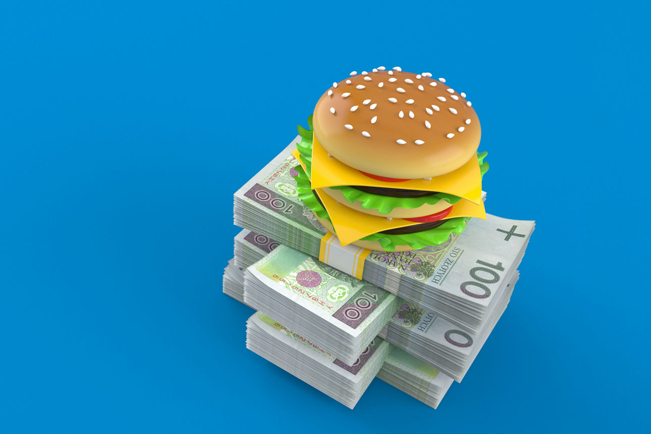 Ein Burger ist auf mehreren Bündeln Gelscheinen gestapelt auf einem hellbauen Hintergrund.